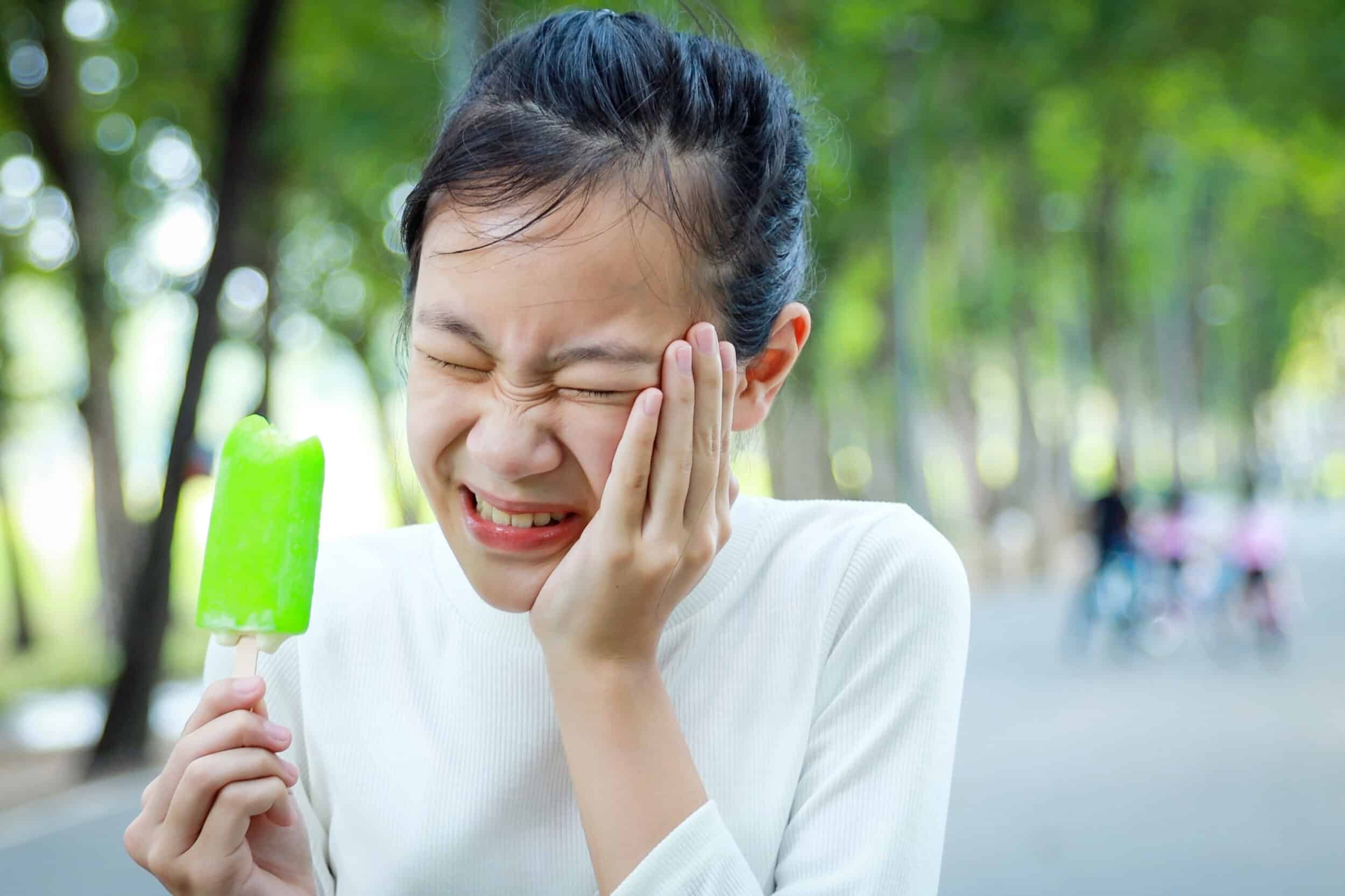 Young girl eating a popsicle and cringing in pain from the cold, in need of dental care
