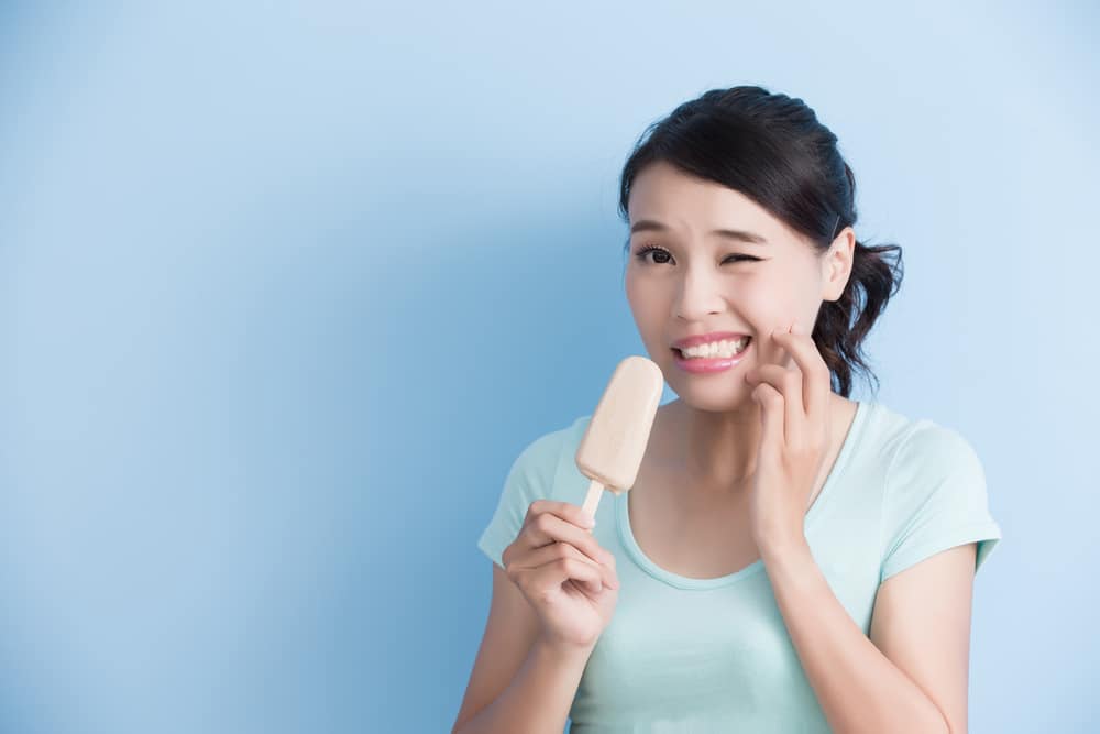 Dentist explaining to a patient what their dental options are, whether keeping the natural tooth is preferred. Young woman struggling with tooth sensitivity and wincing from tooth pain while eating a popsicle.